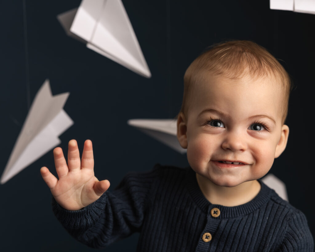 little boy smiling and waving to the camera on a blue backdrop with white paper airplanes 