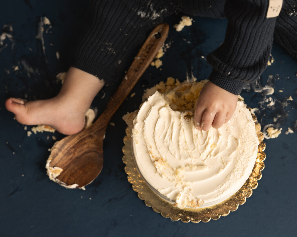 a detail image of a cake smash where we see the child's hands and feet, a large wooden spoon, and the destroyed cake from above