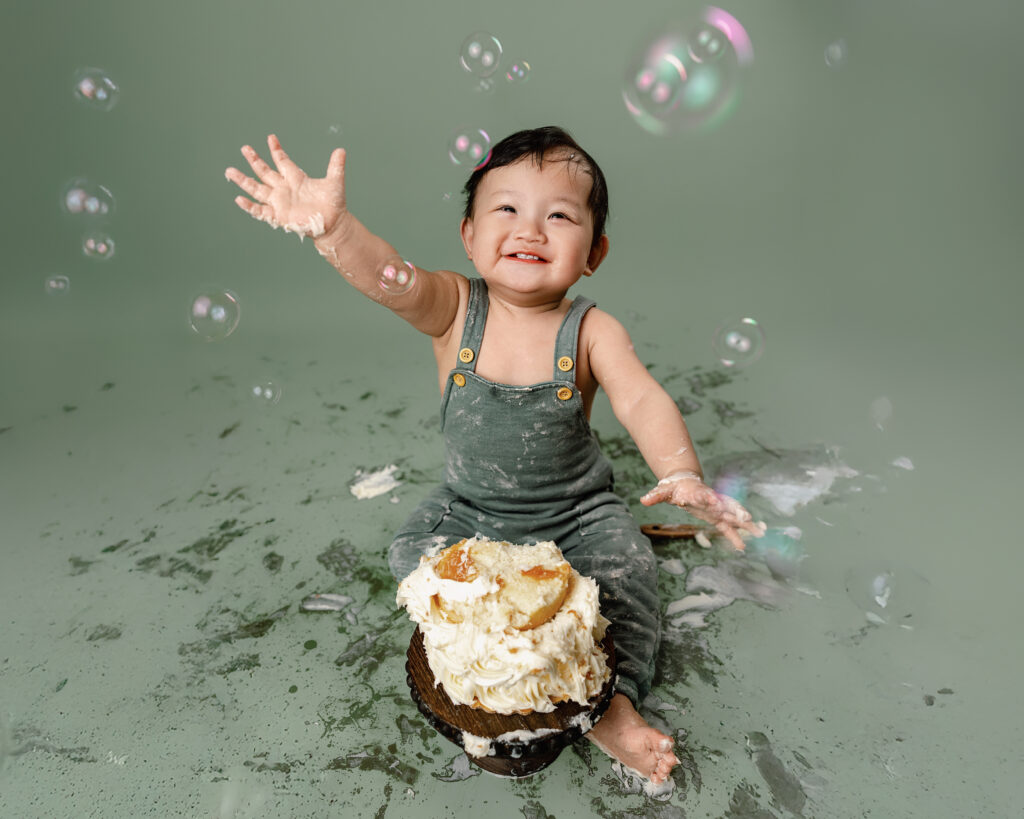 cake smash image of a young boy covered in frosting and smiling at bubbles