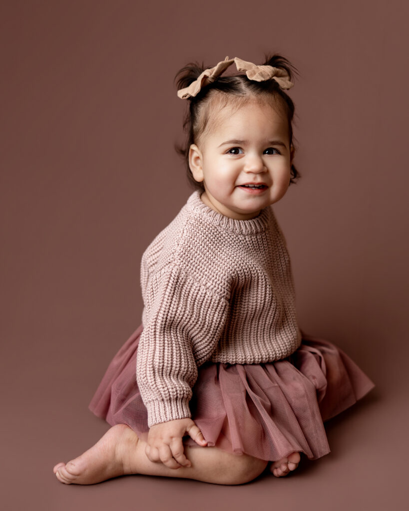 toddler on a colored mauve studio backdrop smiling happily on her knees